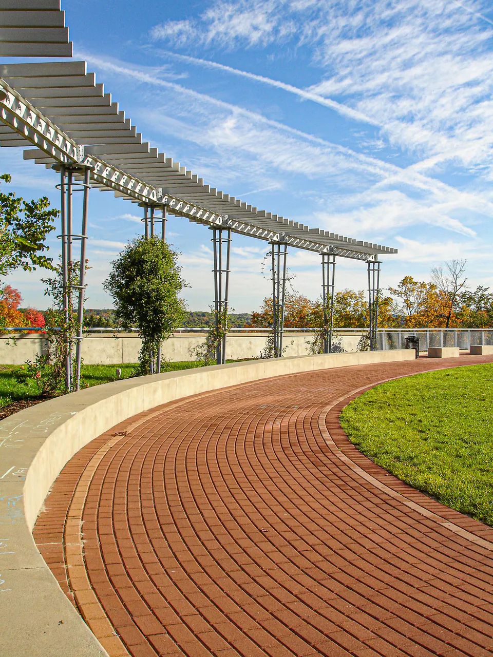 Campus brick walk curving under a white trellis structure beside lawn and young trees